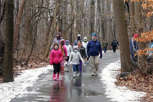A group of children and adults walking on a snowy path through a wooded area, wearing winter clothing.