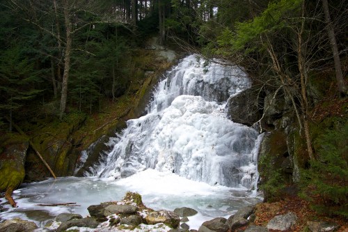 A frozen waterfall cascading over rocks, surrounded by evergreen trees and a snowy landscape.