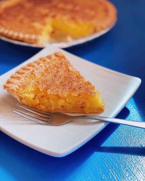 A slice of golden pie on a white plate with a fork, with the whole pie in the background on a blue table.