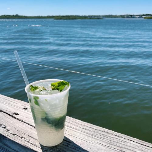 A refreshing mojito in a clear cup sits on a wooden railing by a calm blue water body under a clear sky.