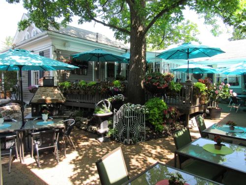 A charming outdoor dining area with turquoise umbrellas, lush greenery, and colorful flowers surrounding a wooden deck.