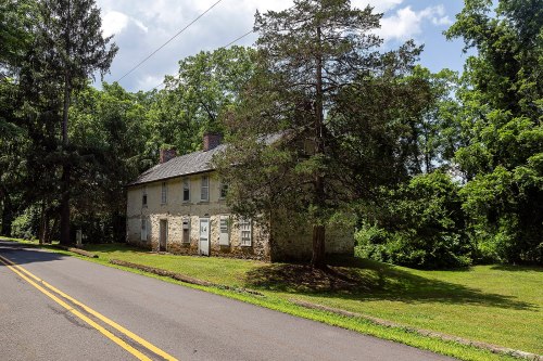 A weathered two-story house surrounded by trees, located near a quiet road under a partly cloudy sky.