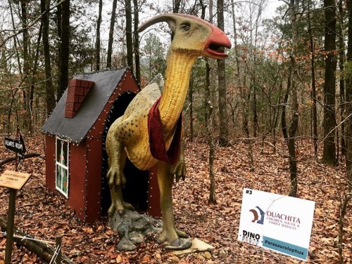 A dinosaur statue wearing a scarf stands next to a small house in a wooded area, with a sign nearby.