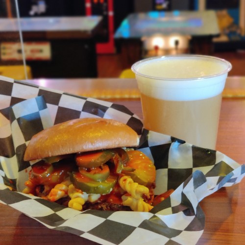A sandwich with pickles and a side of pasta, accompanied by a glass of beer, on a checkered paper plate.