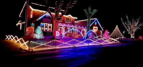 A brightly lit house decorated with colorful Christmas lights, featuring trees and festive displays in the yard.