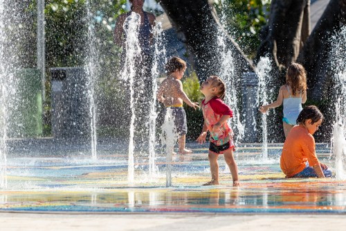 Children play joyfully in a splash pad, surrounded by water fountains on a sunny day.