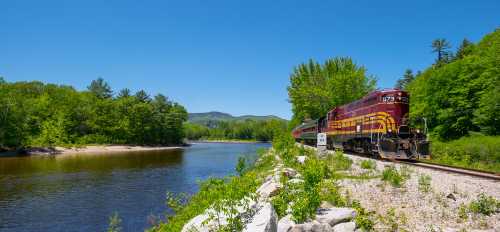 A train travels along a riverbank surrounded by lush green trees and mountains under a clear blue sky.