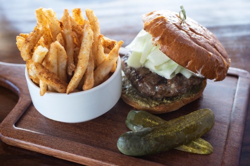 A juicy burger with lettuce on a toasted bun, served with a side of seasoned fries and pickles on a wooden platter.