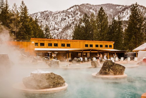 A serene hot spring surrounded by mist, with a wooden building and snow-capped mountains in the background.