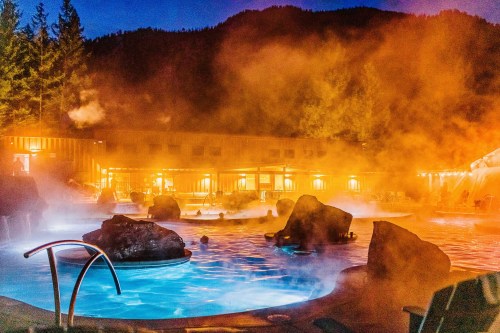 A serene hot springs scene at night, with steam rising over water and glowing lights from nearby buildings.