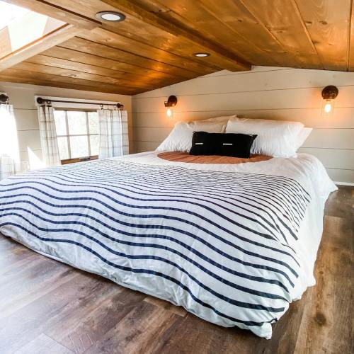 Cozy bedroom with a large bed, wooden ceiling, and natural light from windows, featuring striped bedding and warm lighting.