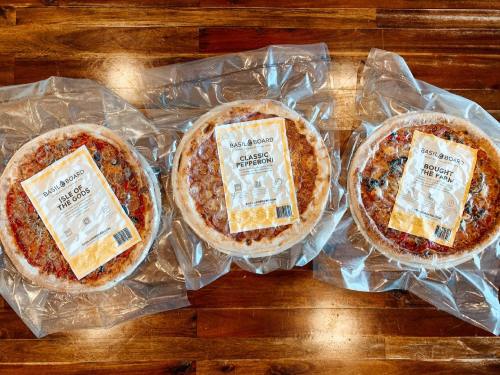 Three packaged pizzas on a wooden surface: one with toppings, classic pepperoni, and a third labeled "Bought the Farm."