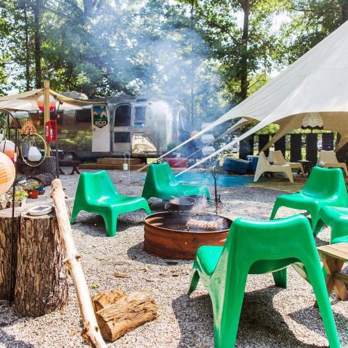 A cozy campsite with green chairs, a fire pit, and a vintage trailer surrounded by trees and outdoor decor.