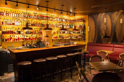 A cozy bar with a wooden counter, shelves lined with various bottles, and warm lighting creating an inviting atmosphere.