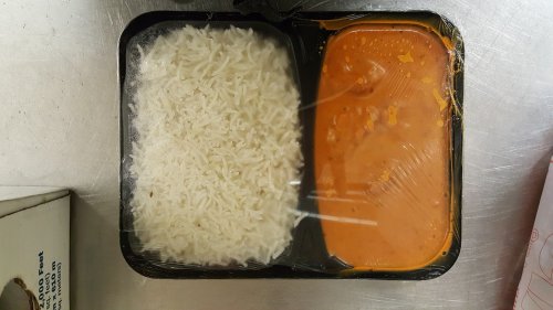 A plastic food container with white rice on one side and orange curry on the other, both covered in plastic wrap.