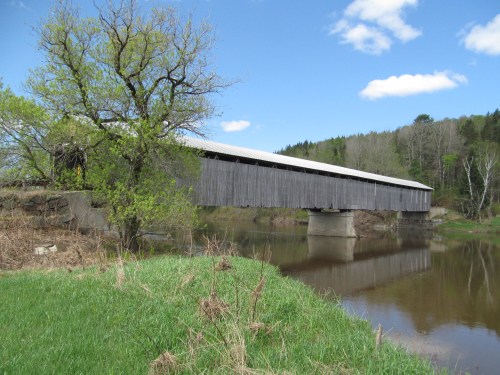 A wooden covered bridge spans a calm river, surrounded by greenery and trees under a blue sky with fluffy clouds.