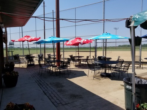Outdoor seating area with tables and colorful umbrellas near a sports field, surrounded by a fence.