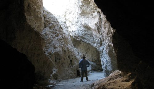 A person stands in a cave, looking up at a bright opening above, surrounded by rocky walls.