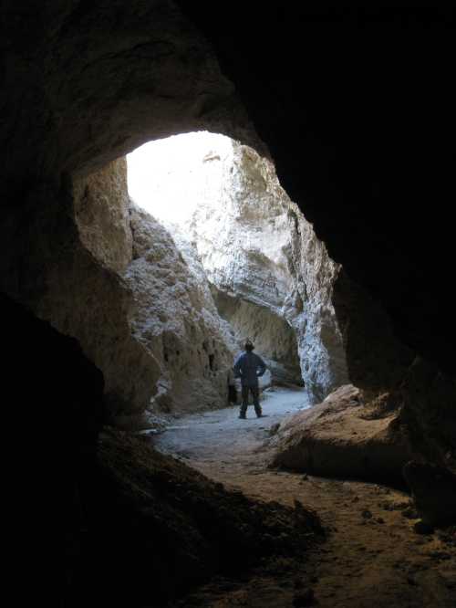 A person stands in a cave, facing a bright opening, surrounded by rocky walls and a sandy floor.