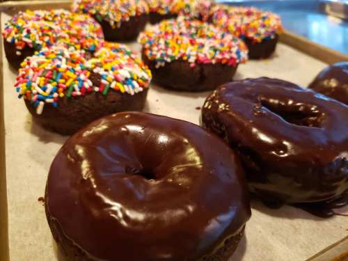 A tray of chocolate donuts, some topped with colorful sprinkles, arranged on parchment paper.