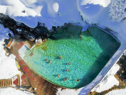 Aerial view of a hot spring pool surrounded by snow, with people relaxing in the warm water.