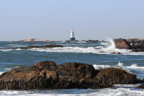 A lighthouse stands on a rocky shore, surrounded by waves and a clear blue sky.