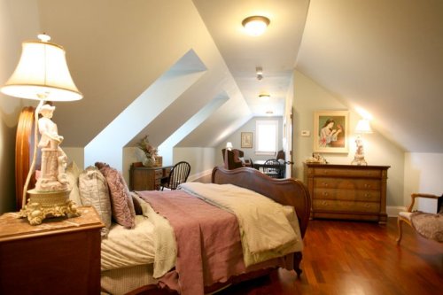 Cozy attic bedroom with sloped ceilings, a wooden bed, dresser, and decorative lamps. Soft colors and vintage decor.