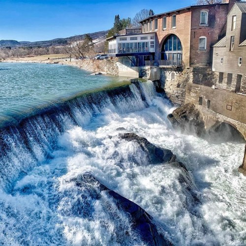 A scenic view of a waterfall cascading near a brick building by the water, with trees and hills in the background.