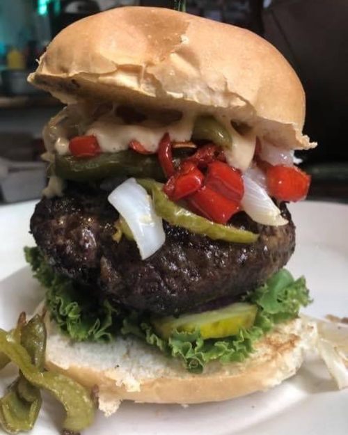 A close-up of a juicy burger with lettuce, pickles, and colorful peppers in a soft bun on a white plate.