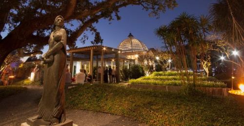 A statue stands in a garden at night, with a glass conservatory and people gathered nearby, illuminated by soft lights.