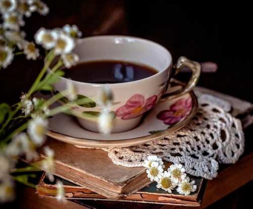 A delicate teacup with flowers sits on stacked books, surrounded by small white flowers and a lace doily.