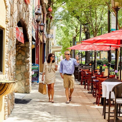 A couple strolls down a sunny street lined with cafes and red umbrellas, enjoying drinks and conversation.