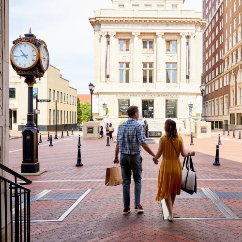 A couple walks hand in hand through a sunny plaza, holding shopping bags, with a historic building in the background.