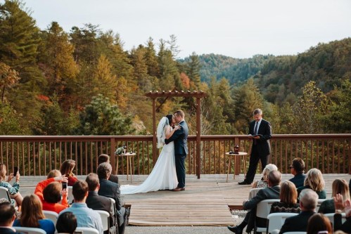 A couple shares a kiss during their outdoor wedding ceremony, surrounded by guests and a scenic mountain backdrop.