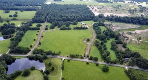 Aerial view of lush green fields, trees, and a winding road near a pond, with a rural landscape in the background.