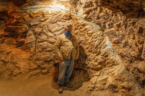 A miner stands against a rocky wall in a dimly lit underground mine, wearing a hard hat and mask.