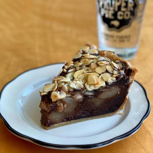 A slice of pie topped with almonds, featuring a chocolate and pear filling, on a white plate with a glass in the background.