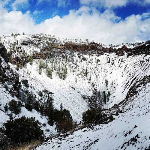 A snowy landscape with a steep, rocky crater surrounded by trees under a blue sky with scattered clouds.