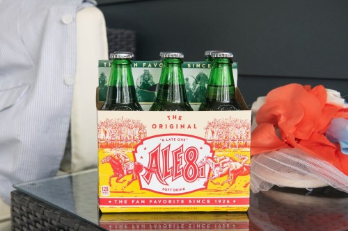 A six-pack of Ale-8 soft drink bottles in a colorful box, sitting on a table with a decorative cloth nearby.
