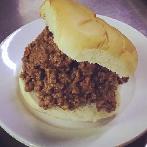 A close-up of a sloppy joe sandwich on a white plate, featuring a soft bun filled with seasoned ground meat.