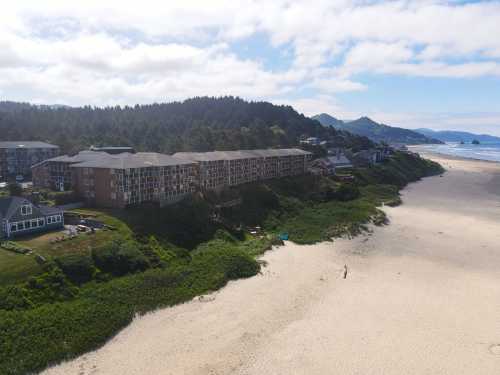 Aerial view of a beach with a hotel, green vegetation, and mountains in the background under a partly cloudy sky.