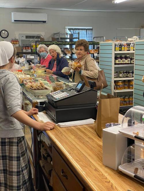 A bakery scene with four women at the counter, chatting and selecting baked goods, while a staff member assists them.