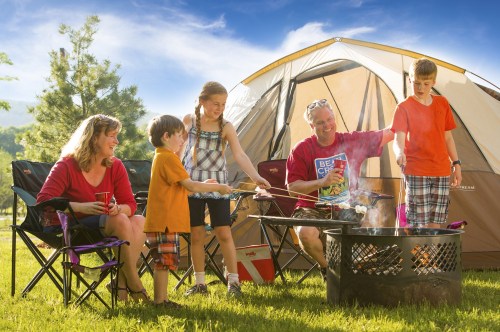 A family enjoys a camping trip, cooking over a fire with a tent in the background and smiles all around.