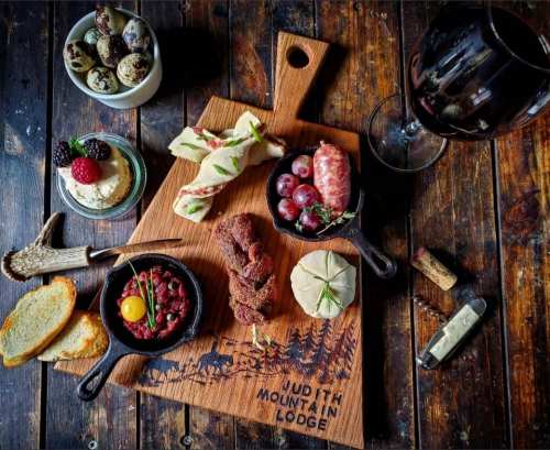 A wooden board with assorted meats, cheeses, fruits, and a glass of red wine, set on a rustic table.