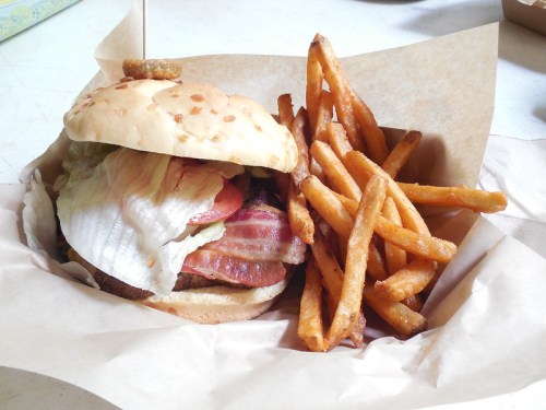 A juicy burger with bacon, lettuce, and tomato, served with a side of crispy sweet potato fries in a paper basket.