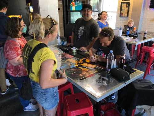 A group of people at a table, with one person signing a poster while others watch and chat in a casual setting.