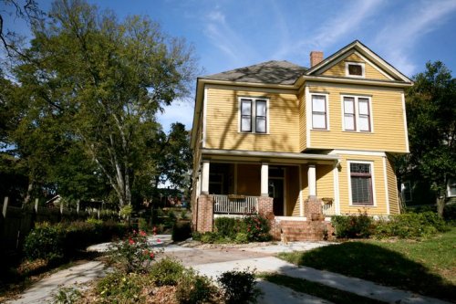 A charming yellow two-story house with a porch, surrounded by greenery and blooming flowers under a blue sky.
