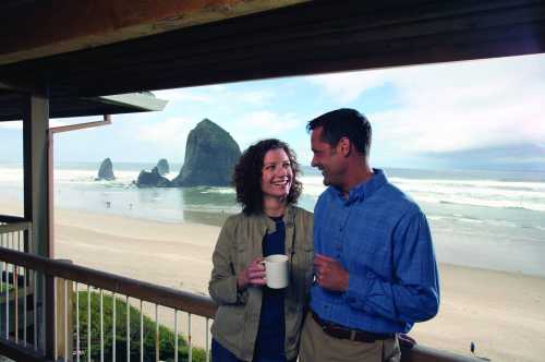 A couple smiles at each other while holding a coffee mug, with a beach and rock formation in the background.