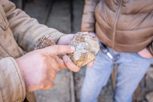 A person points to a rock while holding it, with another person in a brown jacket standing nearby.