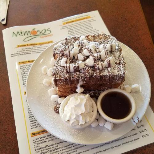 A plate of French toast topped with powdered sugar, mini marshmallows, whipped cream, and a side of syrup. Menu in background.